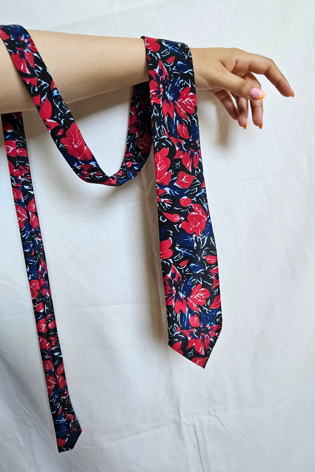 A floral necktie with a navy blue base and red, pink, and white flowers, draped over a person's arm against a white fabric backdrop.