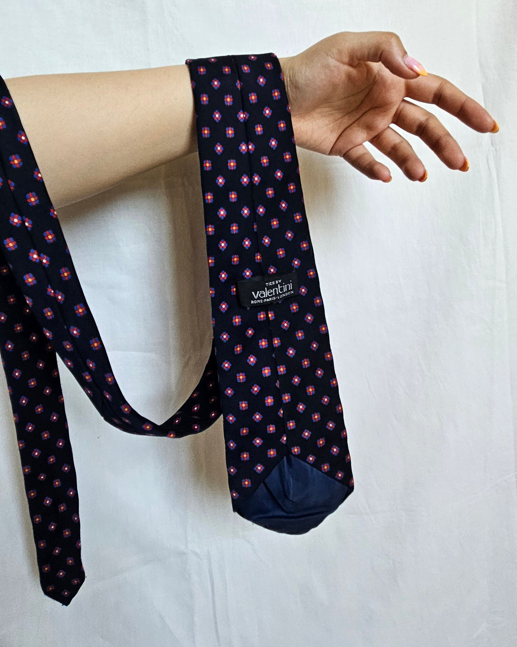 A navy blue necktie with a small pink and white geometric pattern, hanging from a person's hand against a white fabric backdrop.