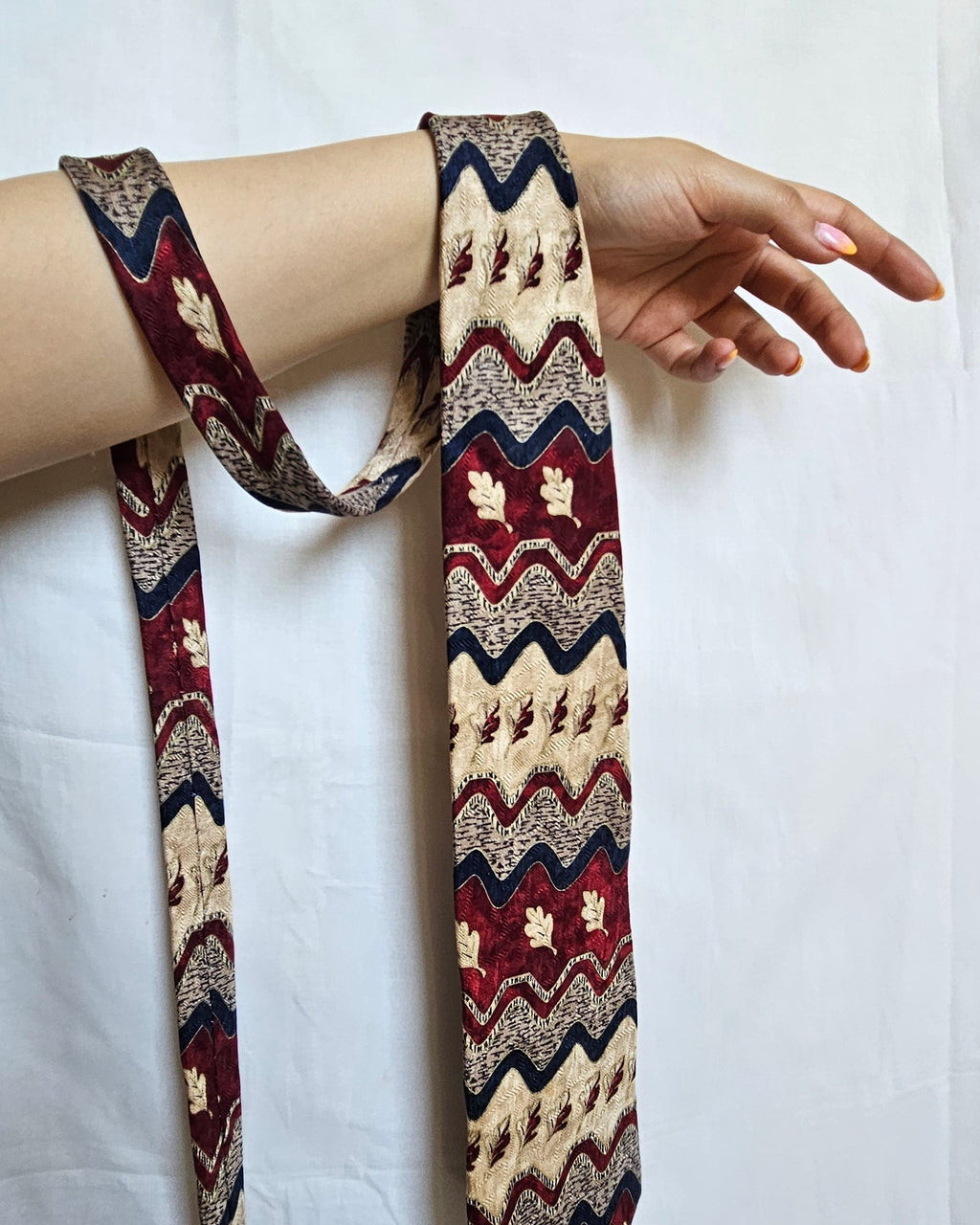 A patterned necktie with a geometric chevron and floral design in burgundy, beige, and navy blue, draped over a person's arm against a white fabric backdrop.