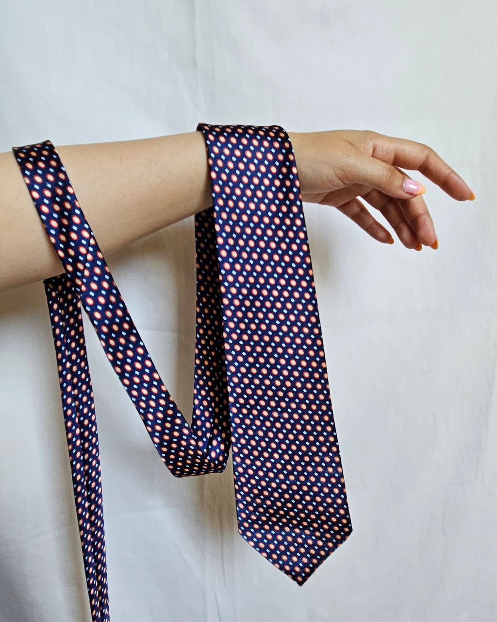 A patterned necktie with a small geometric print in red, yellow, and white on a dark navy base, draped around a person's arm against a white fabric backdrop.