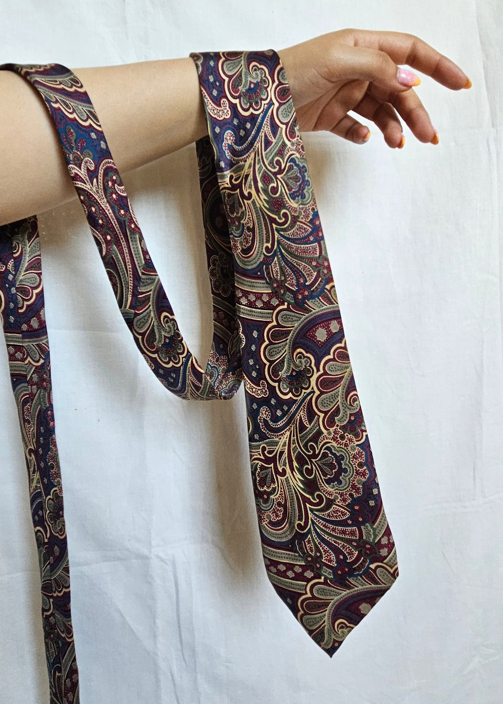 A patterned necktie with an intricate paisley design in purple, gold, and green, draped over a person's arm against a white fabric backdrop.