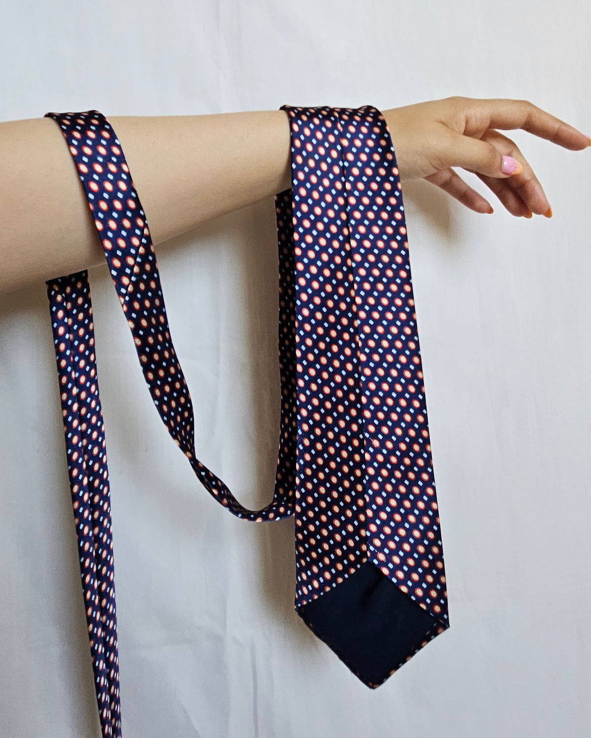 A patterned necktie with a navy blue base and small red, white, and yellow geometric dots, draped over a person's arm against a white fabric backdrop.
