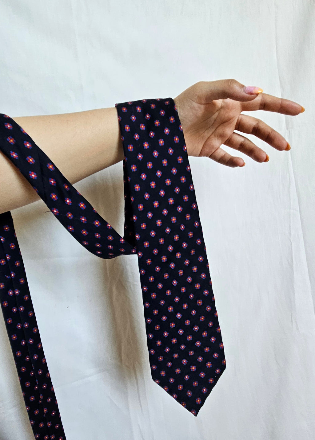 A navy blue necktie with a small pink and green floral pattern, being held by a person's arm and hand against a white fabric backdrop.